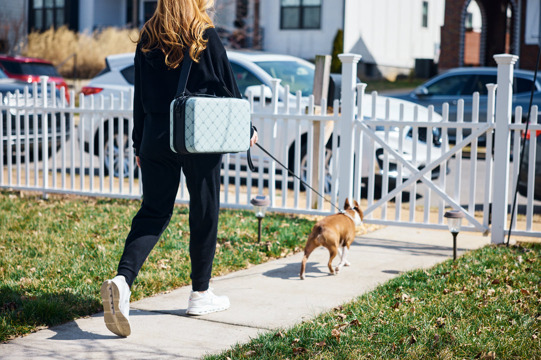 A dog parent walks outdoors carrying a light blue diamond-quilted ZUXI travel case while walking their brown dog on a leash along a sidewalk beside a white picket fence.