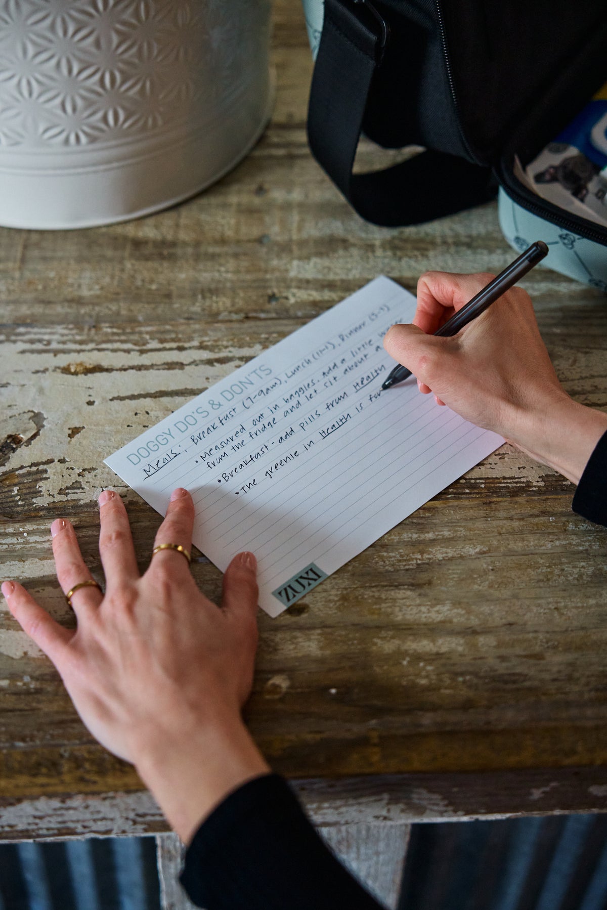 A dog parent's hands handwrite care instructions on a ZUXI "Doggy Do's & Don'ts" card at a weathered wooden table, with a black ZUXI travel case in the background.
