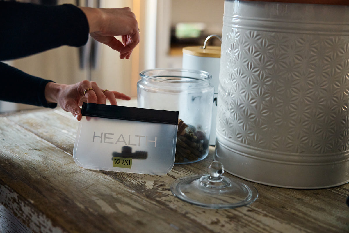 A dog parent drops a treat into a clear ZUXI pouch labeled "HEALTH" on a rustic wooden kitchen counter, next to ceramic canisters.