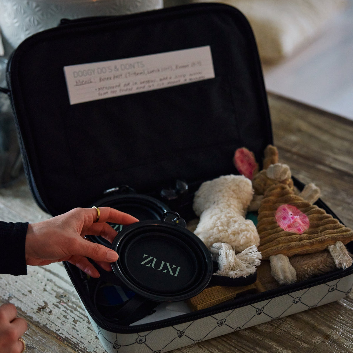 A hand reaches into an open ZUXI travel case packed with plush dog toys, a black collapsible ZUXI bowl, and a handwritten "Doggy Do's & Don'ts" care card, set on a rustic wooden surface.