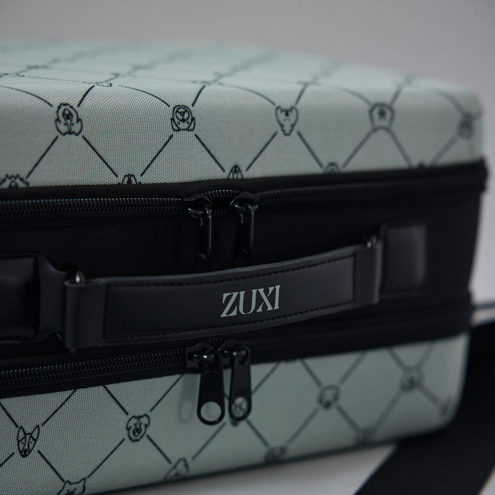 Close-up of the ZUXI travel case in sage green dog-face diamond print fabric, showing the black zippered center band with the ZUXI logo strap.