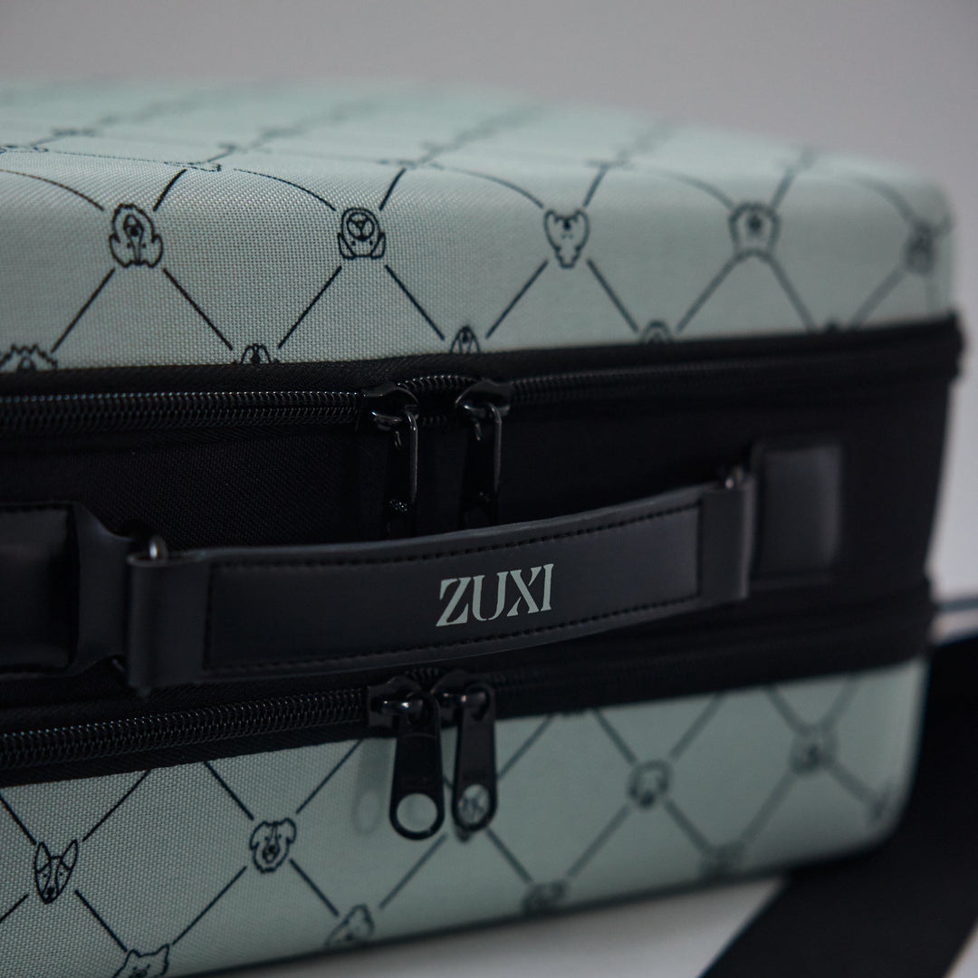 Close-up of the ZUXI travel case in sage green dog-face diamond print fabric, showing the black zippered center band with the ZUXI logo strap.