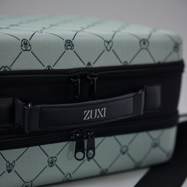 Close-up of the ZUXI travel case in sage green dog-face diamond print fabric, showing the black zippered center band with the ZUXI logo strap.