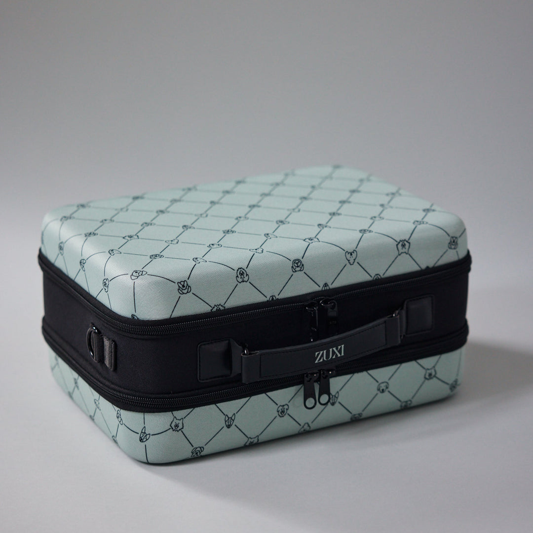 ZUXI travel case in sage green dog-face diamond print fabric with a black zippered middle band and carrying strap bearing the ZUXI logo, shown at an angle against a gray background.