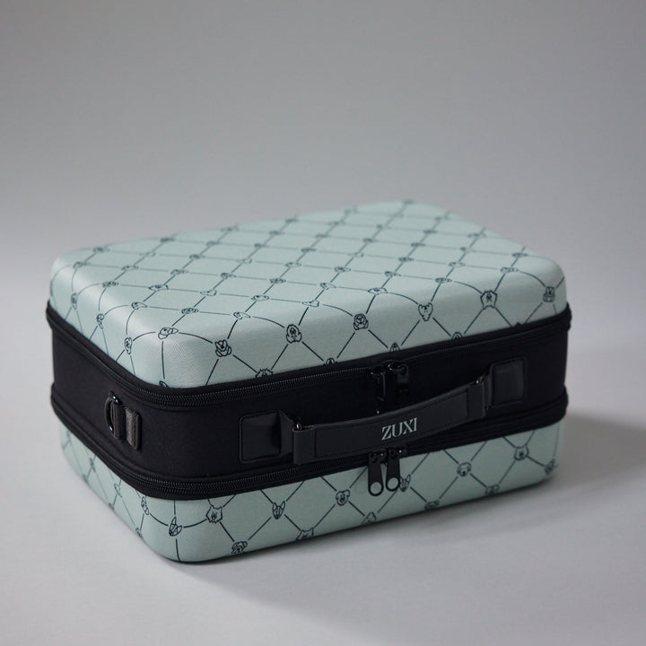 ZUXI travel case in sage green dog-face diamond print fabric with a black zippered middle band and carrying strap bearing the ZUXI logo, shown at an angle against a gray background.