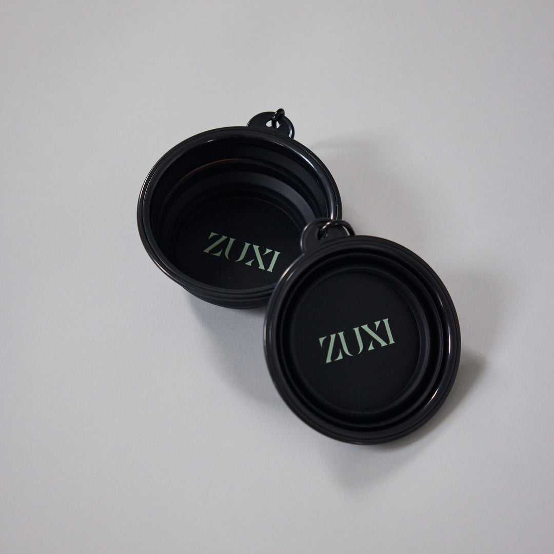 Two black collapsible ZUXI travel bowls with the ZUXI logo printed in sage green inside each bowl, resting on a light gray surface.