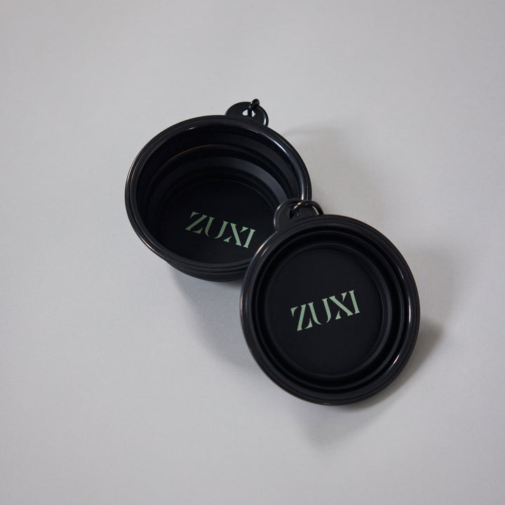 Two black collapsible ZUXI travel bowls with the ZUXI logo printed in sage green inside each bowl, resting on a light gray surface.