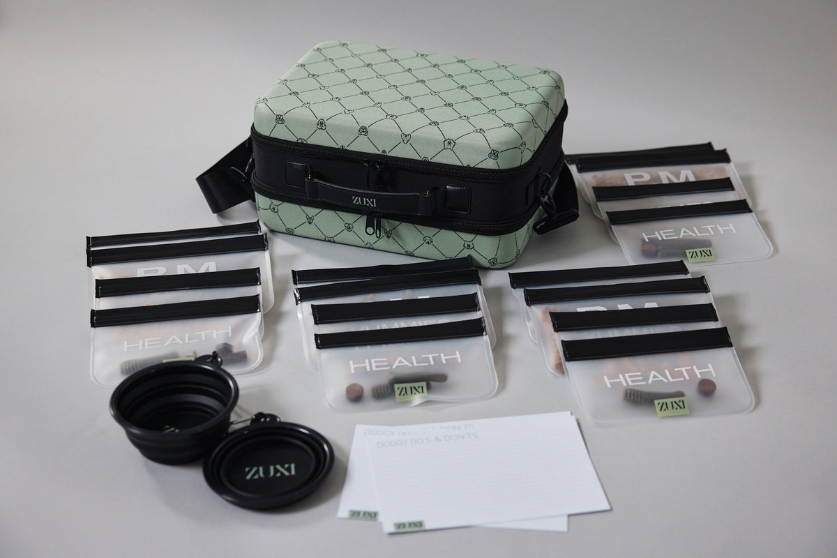 ZUXI dog travel case in sage green diamond-quilted fabric with black trim, surrounded by multiple clear zippered pouches labeled PM and HEALTH, two black collapsible travel bowls, and a printed care instruction card laid out on a white background.
